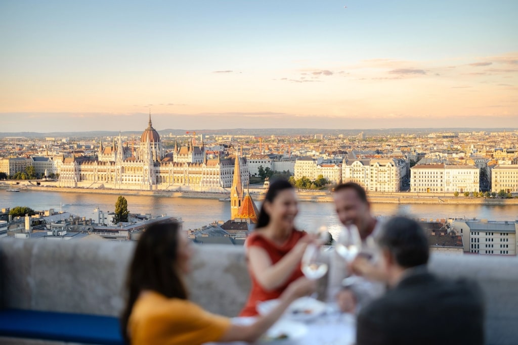 Friends clink glasses during a rooftop dinner, enjoying sweeping golden-hour views of the Danube River and the Hungarian Parliament Building.