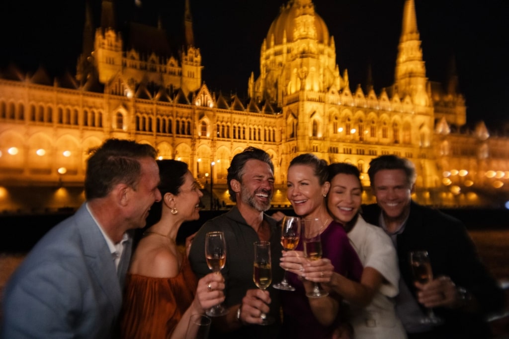 A group of friends toasts with wine during an evening Danube cruise, illuminated by the golden glow of Budapest's grand Parliament Building.
