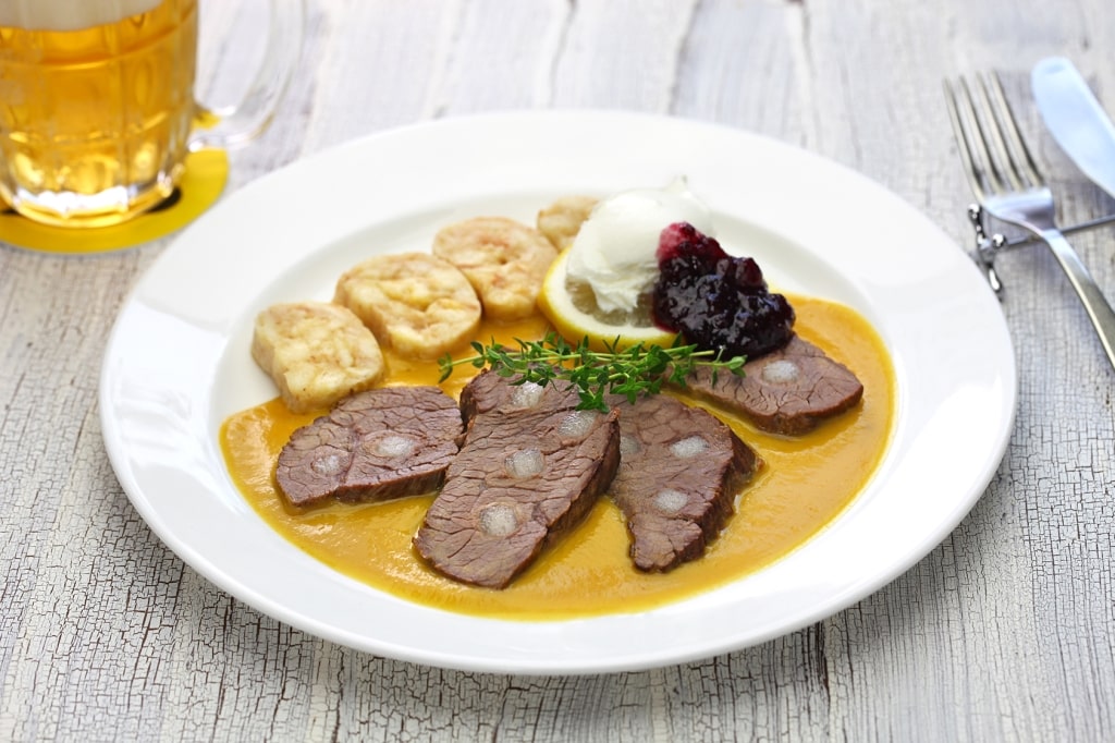 Tender slices of beef in a rich, creamy root-vegetable sauce are paired with soft bread dumplings, cranberries, and a cold glass of golden Czech beer.