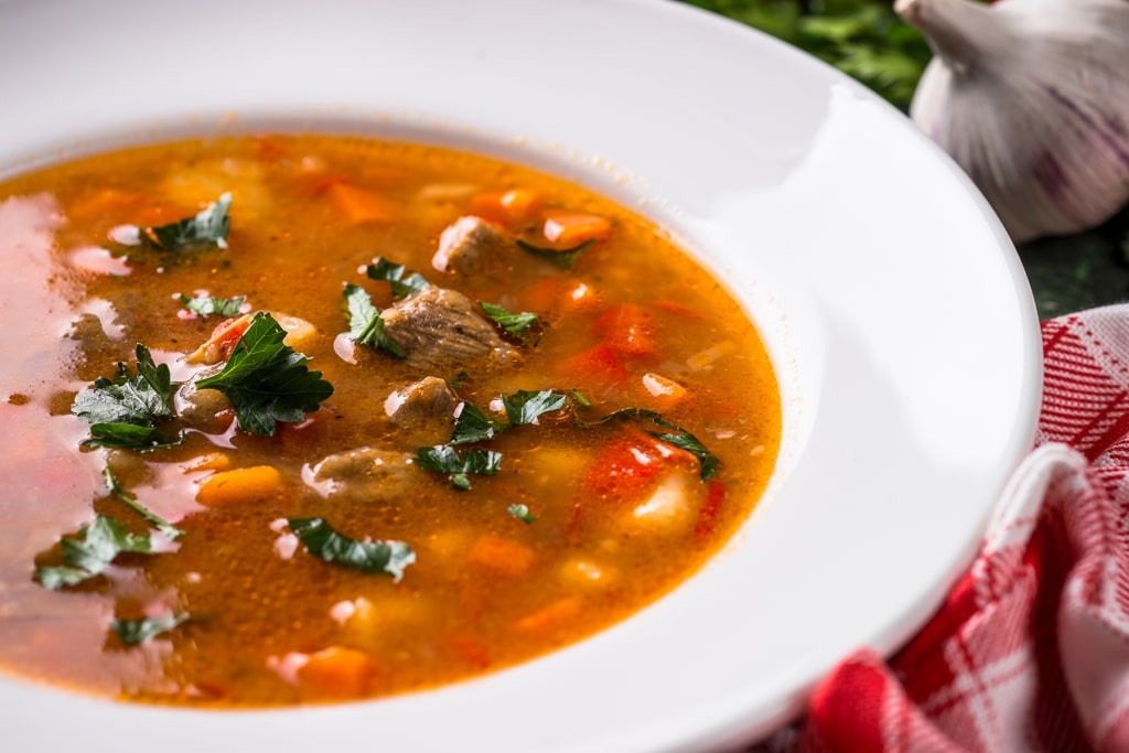A white bowl of hearty Hungarian goulash soup brims with tender beef, diced vegetables, and fresh parsley beside a classic checkered napkin.