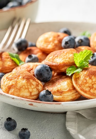 Fluffy golden Dutch poffertjes scattered with plump blueberries and fresh mint leaves are served as a sweet treat on a ceramic plate.