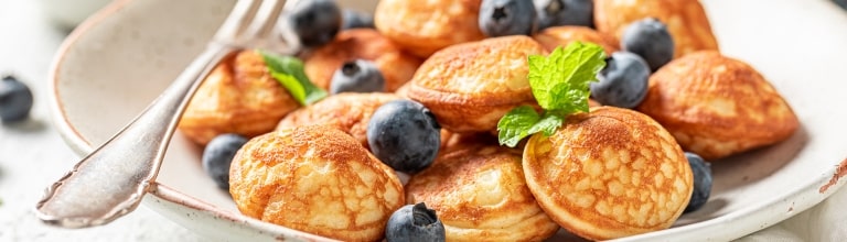 Fluffy golden Dutch poffertjes scattered with plump blueberries and fresh mint leaves are served as a sweet treat on a ceramic plate.