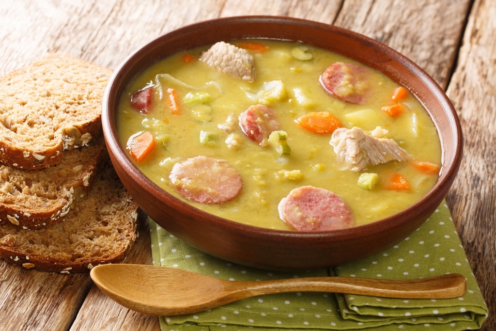 A rustic bowl of thick Dutch split pea soup filled with sliced sausage and vegetables rests alongside slices of dense rye bread.
