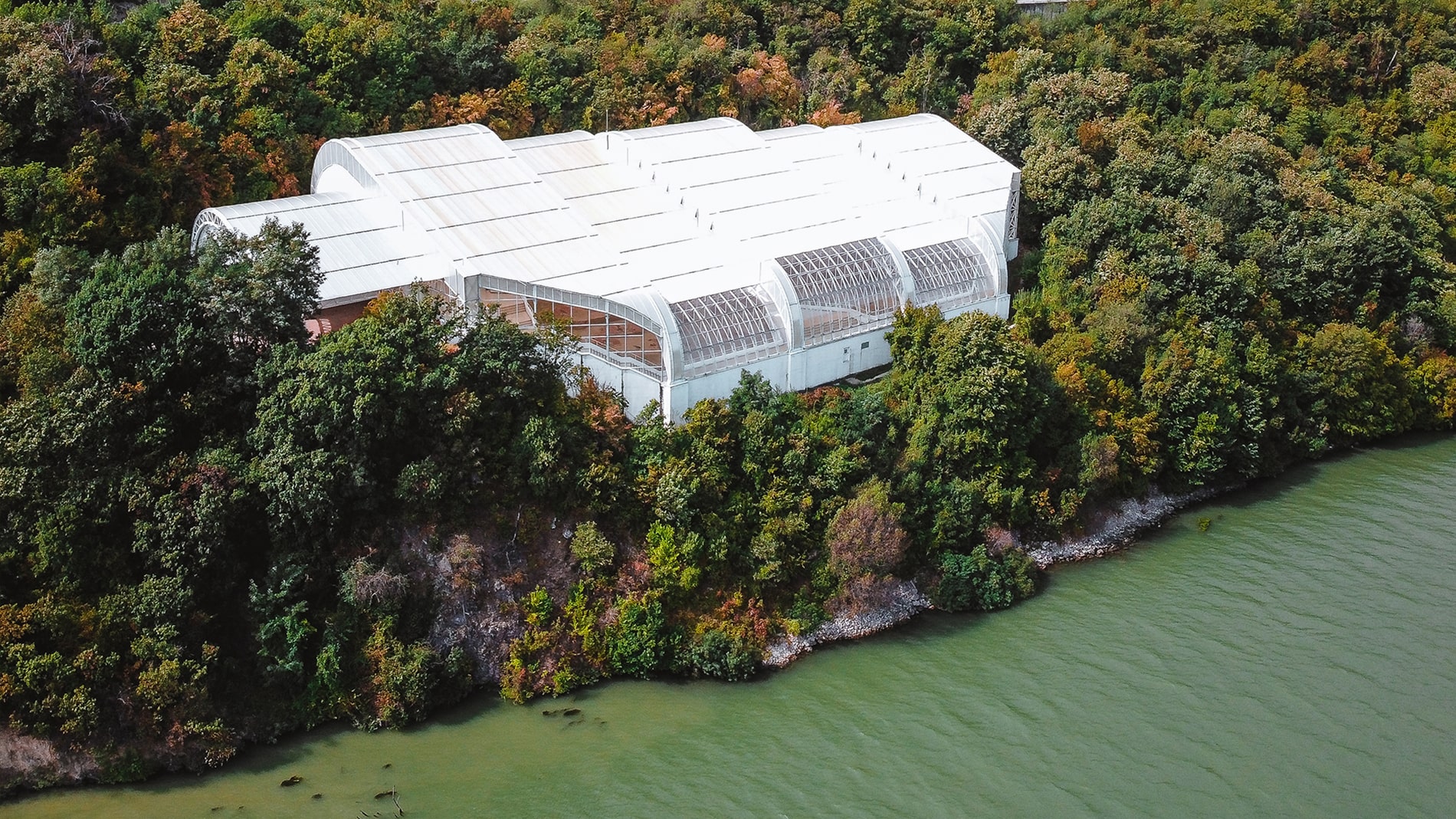The modern white protective enclosure of the Lepenski Vir archaeological site rests on the lush banks of the Danube River beneath steep, rocky slopes