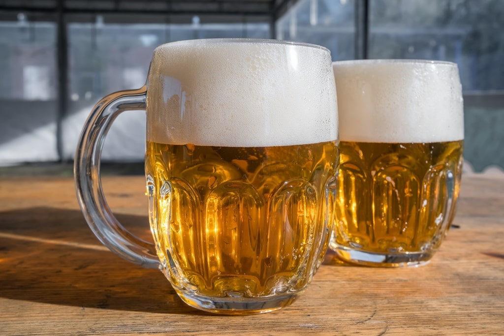 Sunlight illuminates two heavy glass mugs of crisp, golden Czech lager topped with thick, creamy foam on a rustic wooden table.