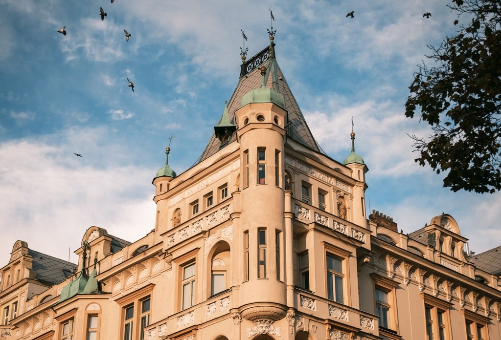 A flock of birds swoops through the sky above the ornate turrets of an elegant historic building in Prague's Žižkov district.