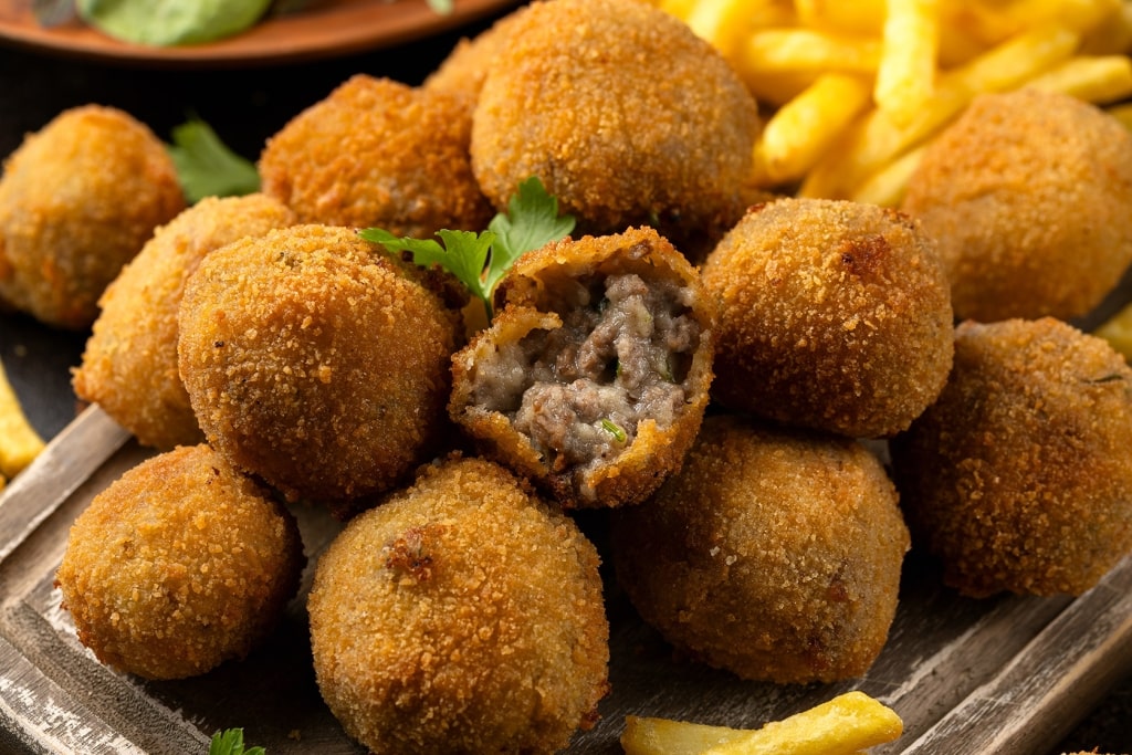 Crispy golden-brown Dutch bitterballen break open to reveal a rich, savory beef filling, served with french fries on a wooden board.