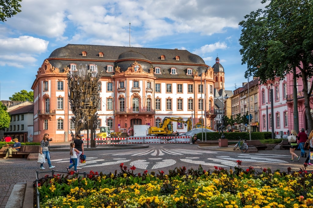 The grand Baroque facade of the Osteiner Hof frames Schillerplatz in Mainz, where vibrant flowerbeds and public fountains anchor a bustling Rhine river cruise stop.