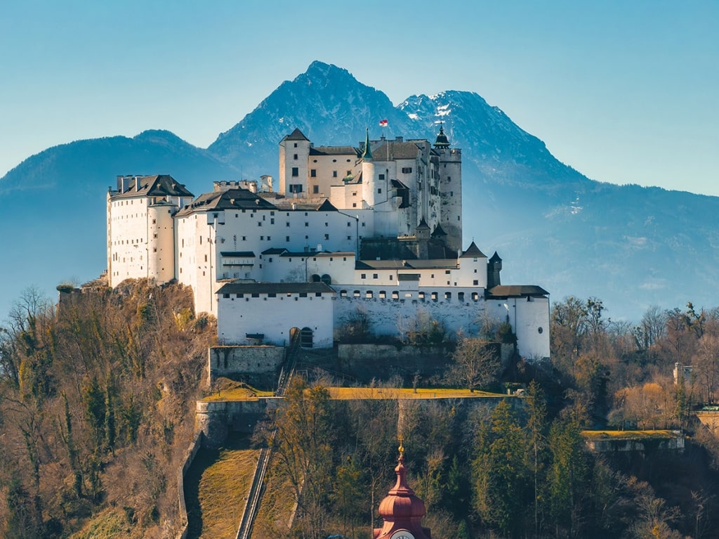 Hohensalzburg Fortress atop a hill in Salzburg, Austria, with white walls, medieval architecture, lush greenery, and the Alps under a clear blue sky.