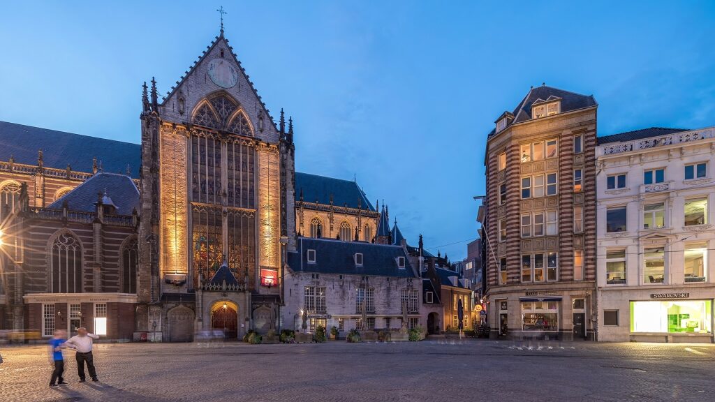 The illuminated facade of the Gothic Nieuwe Kerk anchors Dam Square in central Amsterdam at twilight.