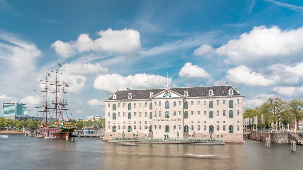 The historic three-masted replica ship Amsterdam sits docked beside the grand National Maritime Museum under a bright, cloud-dappled sky.