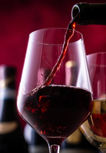 A stream of rich red wine pours gracefully into a clear glass beside a glass of white wine, set against a dramatic, ruby-lit backdrop.