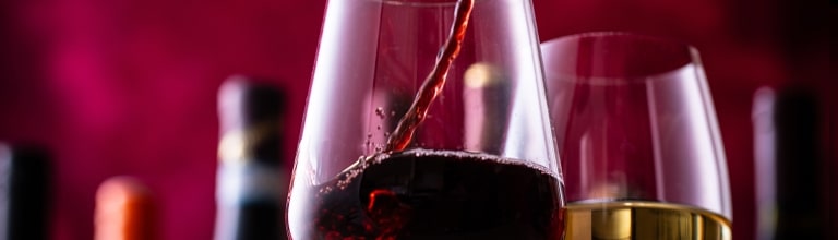 A stream of rich red wine pours gracefully into a clear glass beside a glass of white wine, set against a dramatic, ruby-lit backdrop.