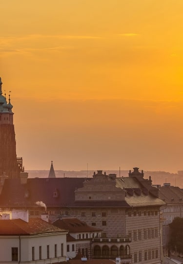 The spires of St. Vitus Cathedral rise against a glowing golden sunrise over Prague.