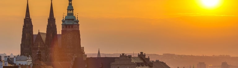 The spires of St. Vitus Cathedral rise against a glowing golden sunrise over Prague.