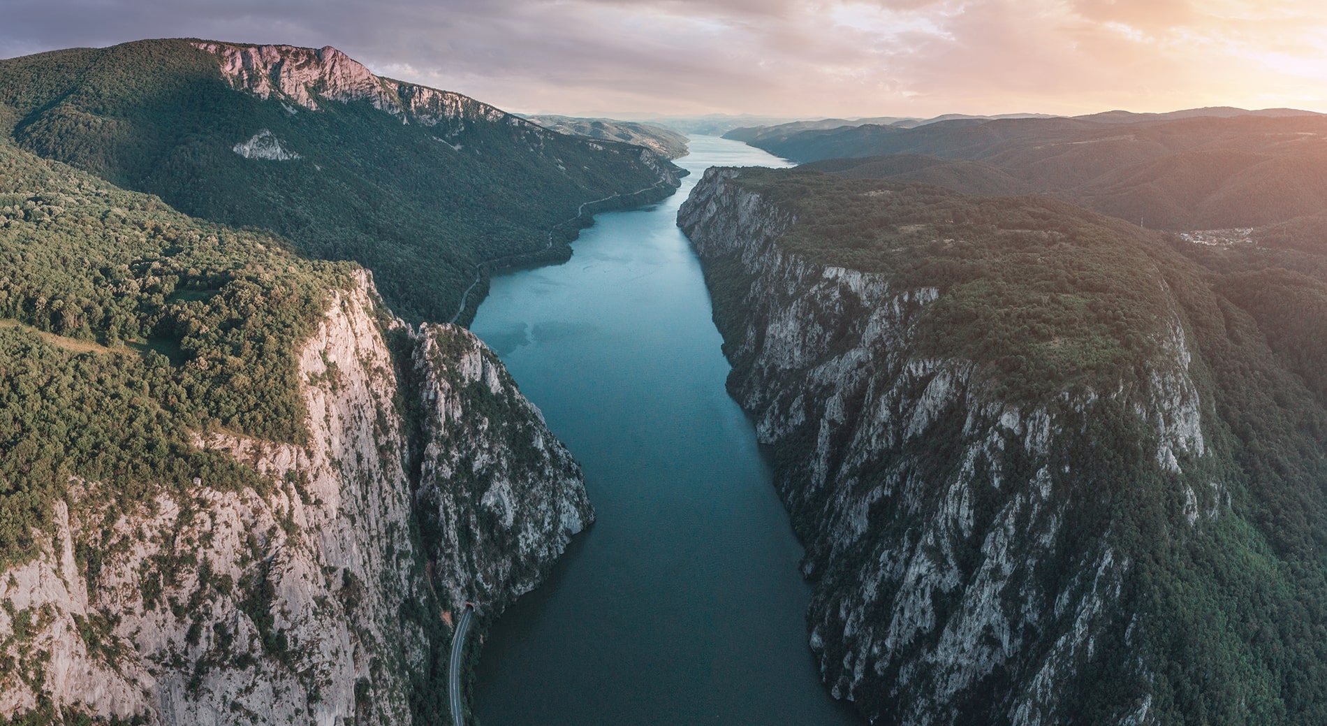 Aerial view captures the Danube River winding through the dramatic Iron Gates gorge, flanked by sheer limestone cliffs and lush green forests under a soft, golden sky