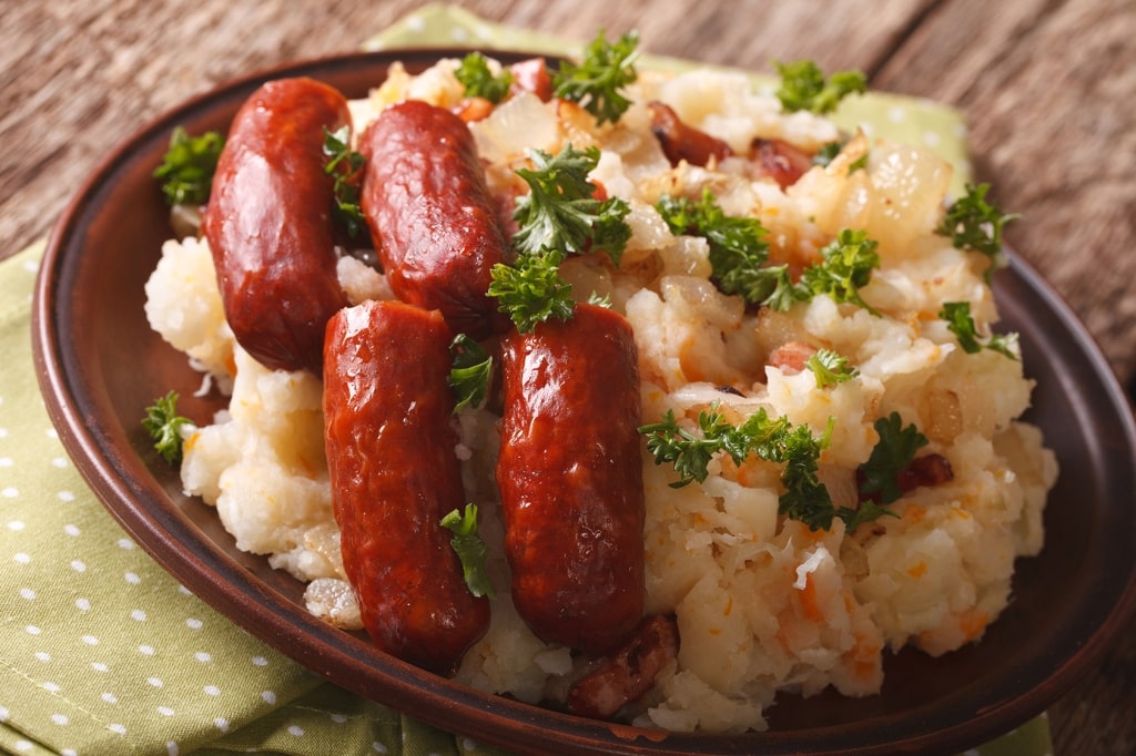 Seared sausages crown a plate of traditional stamppot mashed potatoes mixed with cabbage and carrots, garnished with fresh parsley.