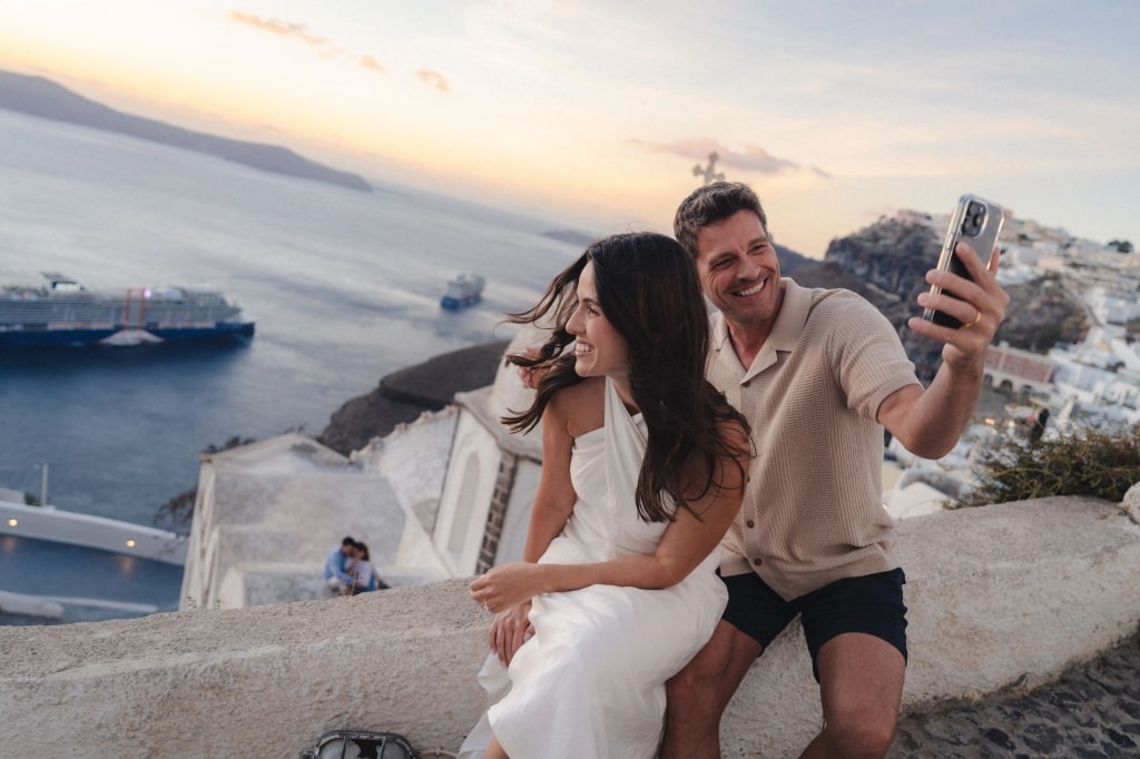 A smiling couple captures a selfie overlooking Santorini's whitewashed cliffs and blue waters, protected by cruise travel insurance on their Celebrity Cruises getaway.