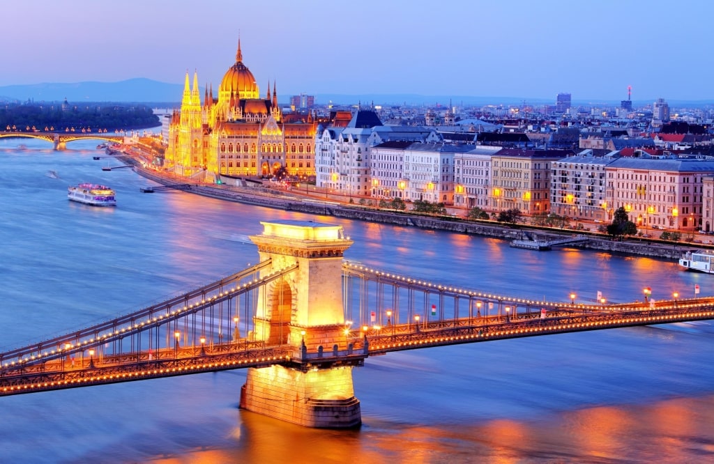The illuminated Széchenyi Chain Bridge spans the Danube River leading toward the glowing Hungarian Parliament Building at twilight.