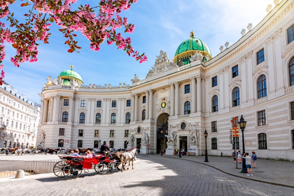 A horse-drawn carriage passes through St. Michael's Square (Michaelerplatz) framed by pink spring blossoms in front of the Hofburg Palace in Vienna.