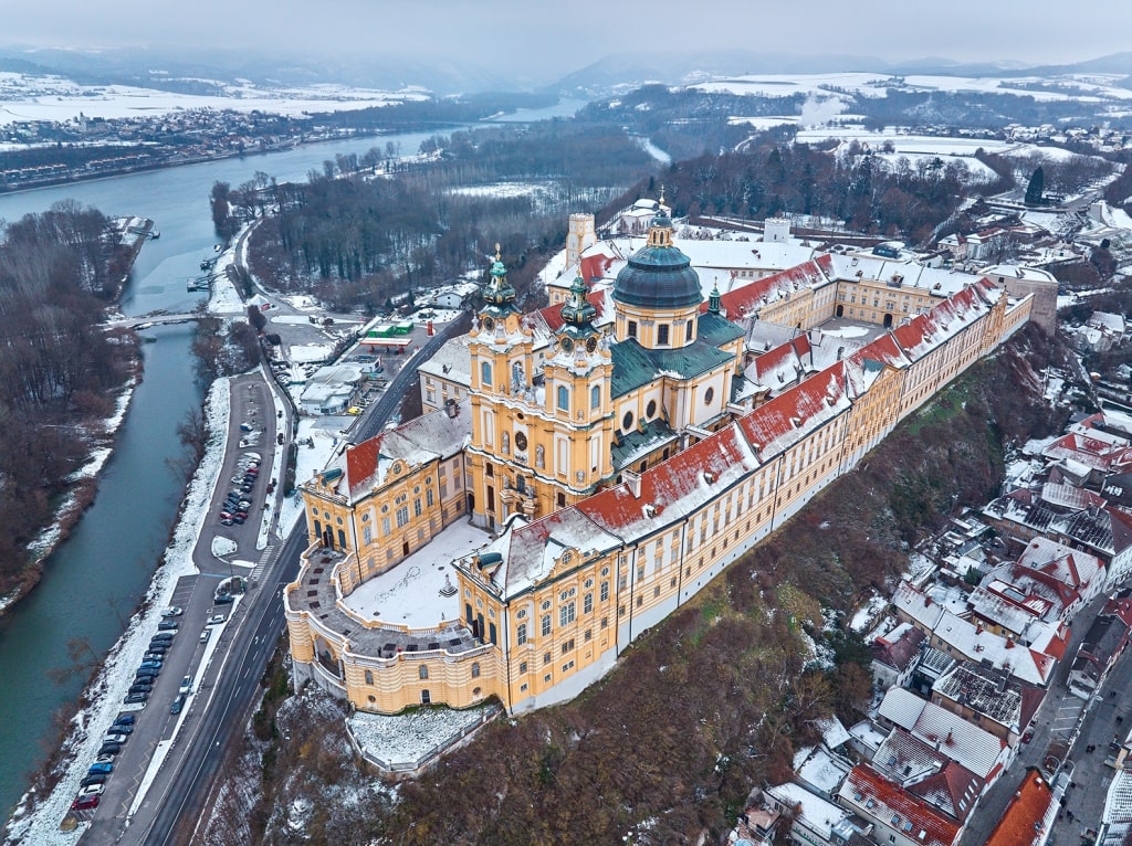 A blanket of winter snow covers the historic town and the towering Benedictine monastery from an aerial perspective.