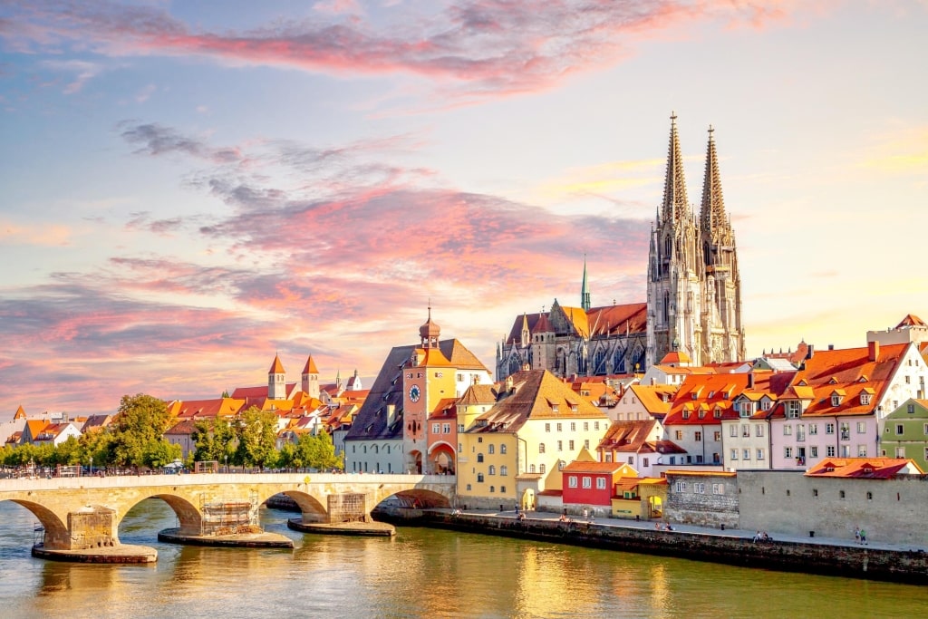 Sunset over the historic Old Town of Regensburg, featuring the medieval Stone Bridge spanning the Danube River and the twin spires of Regensburg Cathedral.