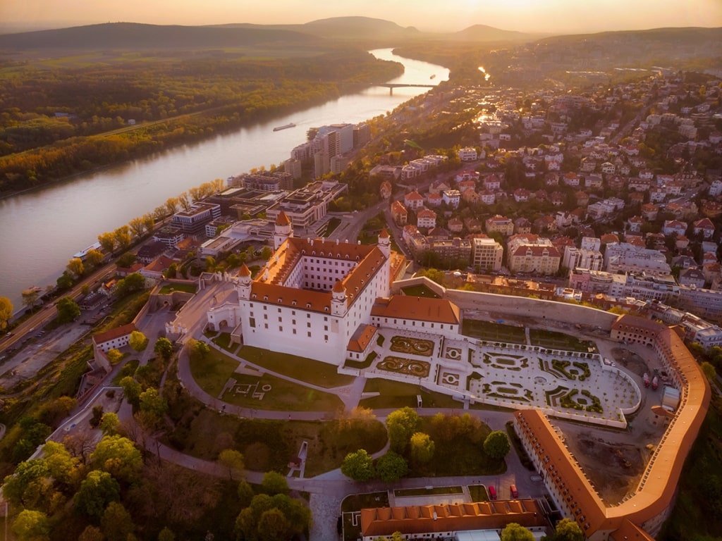 A golden hour aerial view of Bratislava Castle (Bratislavský hrad), showcasing its central courtyard, Baroque garden, and the Danube River winding through the landscape.