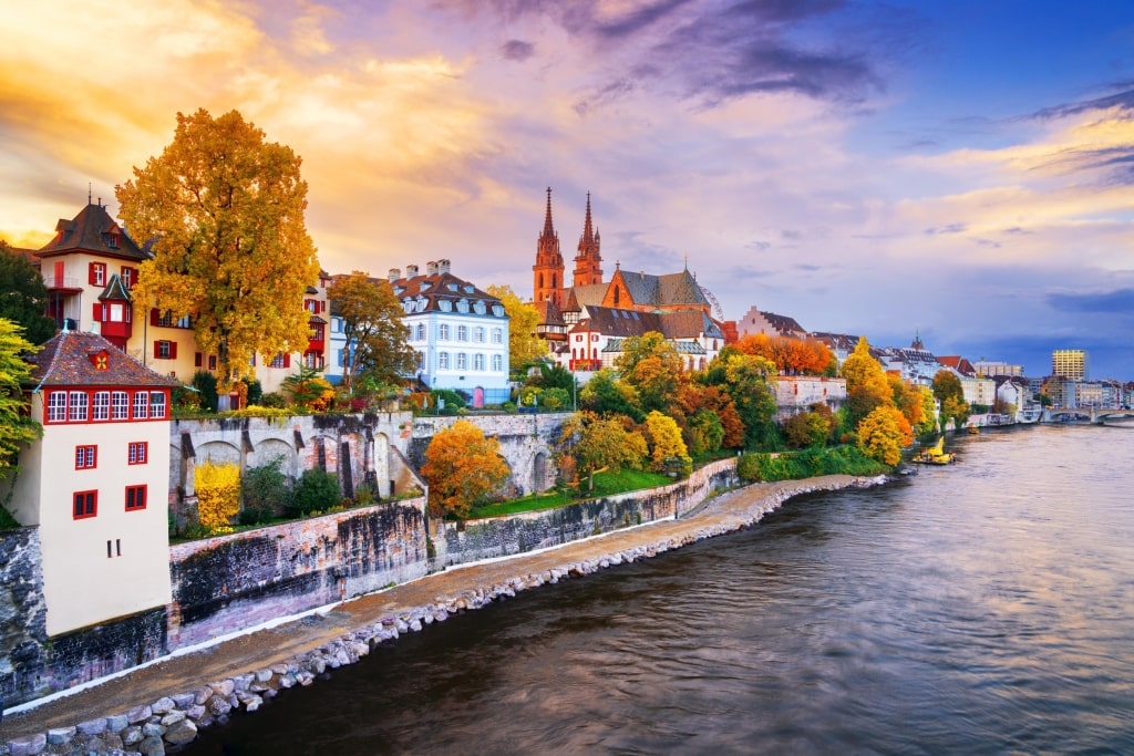 Autumn colors line the banks of the Rhine River, overlooked by the red sandstone spires of the Basel Minster cathedral at sunset.