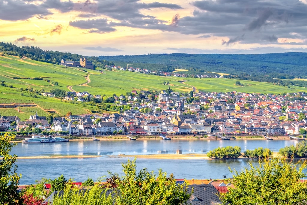A panoramic view of the picturesque town of Rüdesheim set against rolling vineyard hills along the banks of the Rhine River.