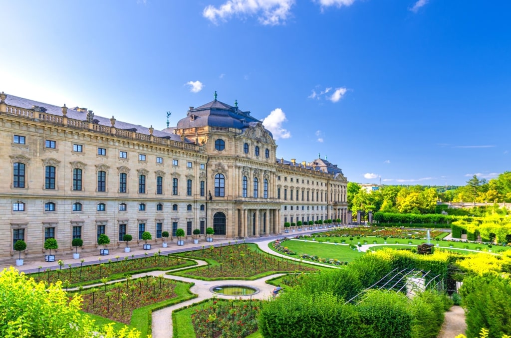 The grand Baroque facade of the Würzburg Residence and its meticulously manicured Court Garden (Hofgarten) under a clear blue sky.