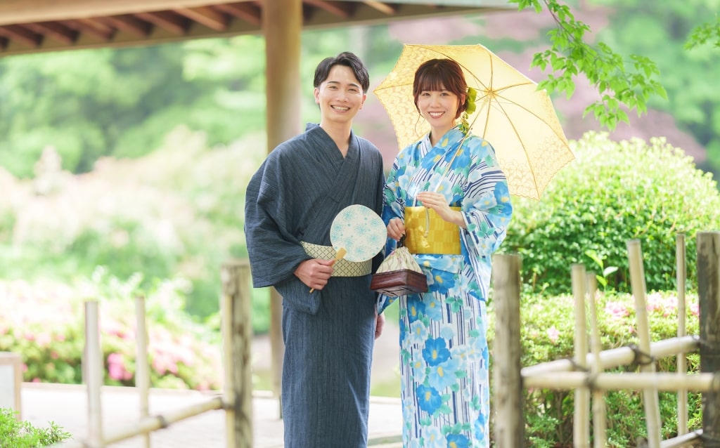 A couple dressed in traditional summer yukata holding a paper fan and parasol while strolling through a lush Japanese garden.