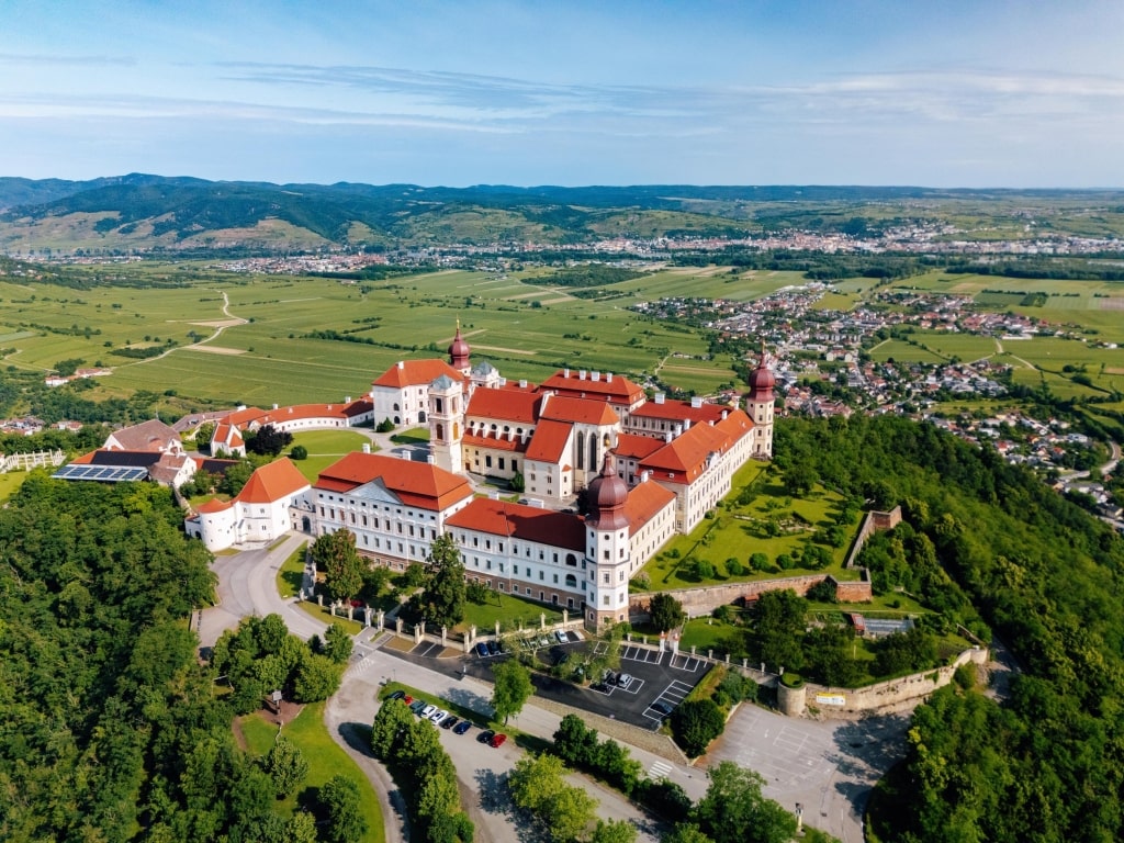 An aerial view captures the majestic, palace-like complex of Göttweig Abbey perched atop a hill surrounded by lush green landscapes.