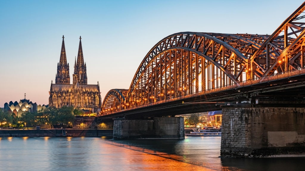 Dusk over the Rhine River featuring the illuminated Hohenzollern Bridge leading toward the Gothic twin spires of Cologne Cathedral.