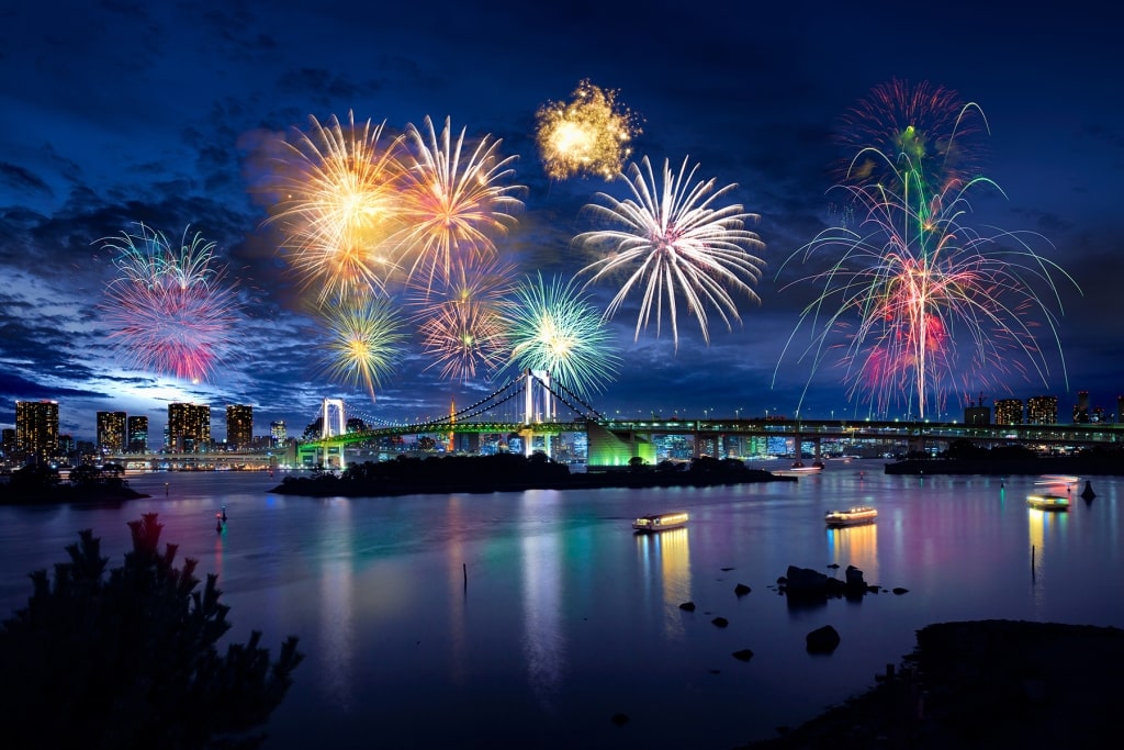 Colorful fireworks illuminate the night sky above the Rainbow Bridge and Tokyo Bay, framed by the distant city skyline during a Hanabi Taikai festival.