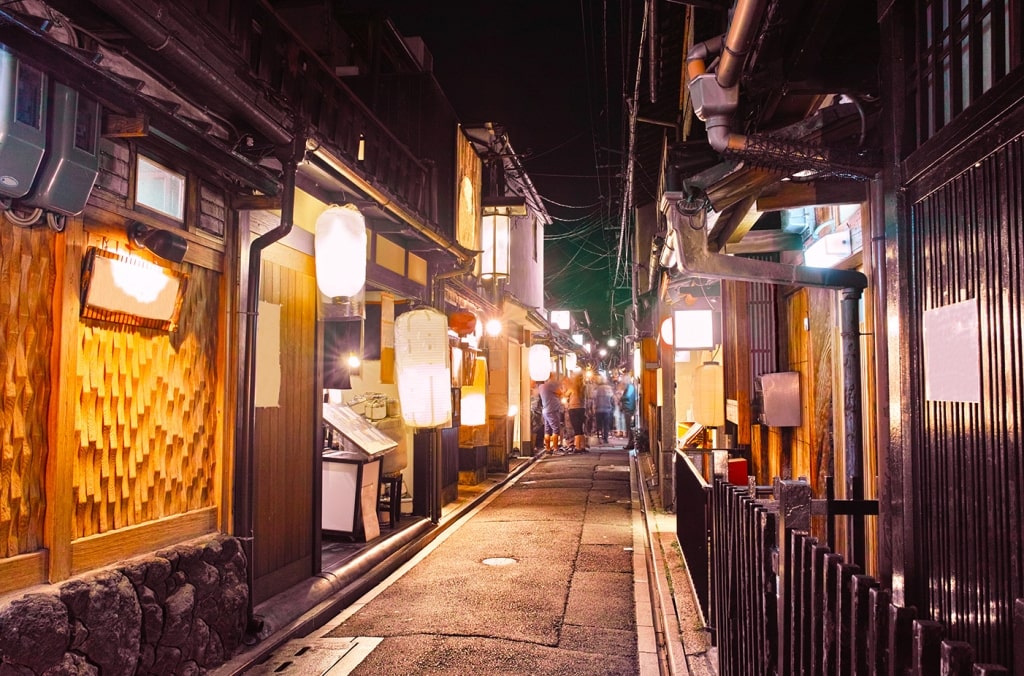 Lanterns illuminate a narrow atmospheric alleyway of traditional wooden restaurants and teahouses in Pontocho Alley at night.
